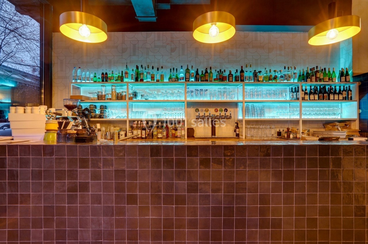 Bar moderne avec comptoir carrelé sombre, étagères éclairées affichant bouteilles et verres, et suspensions lumineuses…