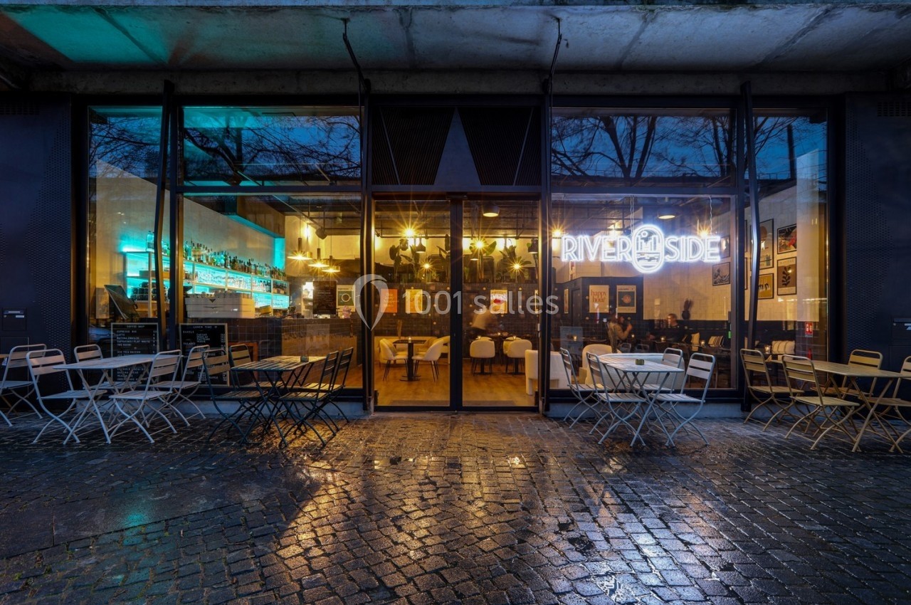Façade vitrée d'un café-restaurant avec enseigne lumineuse, tables extérieures et sol pavé humide.