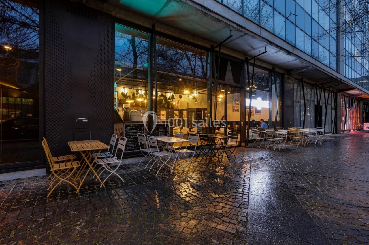Terrasse d'un café avec tables et chaises en métal, devant une façade vitrée, sur une rue pavée mouillée.