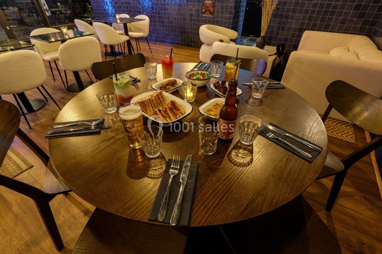 Table ronde en bois dressée avec des plats variés, boissons et couverts, dans un restaurant à l'ambiance chaleureuse.