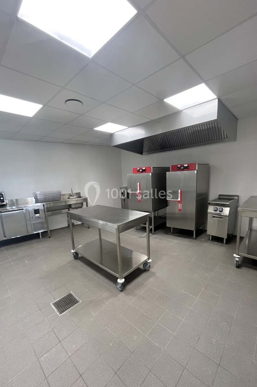 Cuisine professionnelle équipée avec plan de travail inox, fours, hotte, micro-ondes et sol carrelé gris.