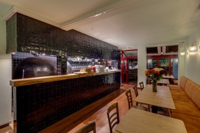 Bar avec comptoir en carrelage sombre, tabourets en bois, étagères avec bouteilles et verres, éclairage suspendu.