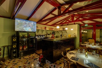 Bar avec comptoir en carrelage sombre, tabourets en bois, étagères avec bouteilles et verres, éclairage suspendu.