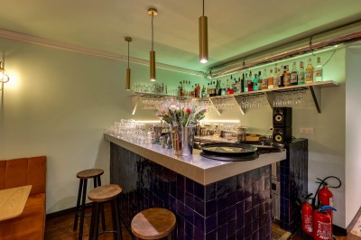 Bar avec comptoir en carrelage sombre, tabourets en bois, étagères avec bouteilles et verres, éclairage suspendu.