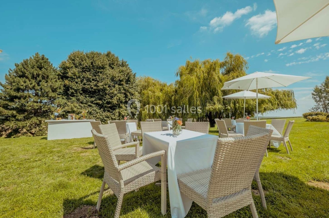 Tables et chaises en rotin disposées sur une pelouse, entourées d'arbres, avec des parasols blancs par temps ensoleillé.