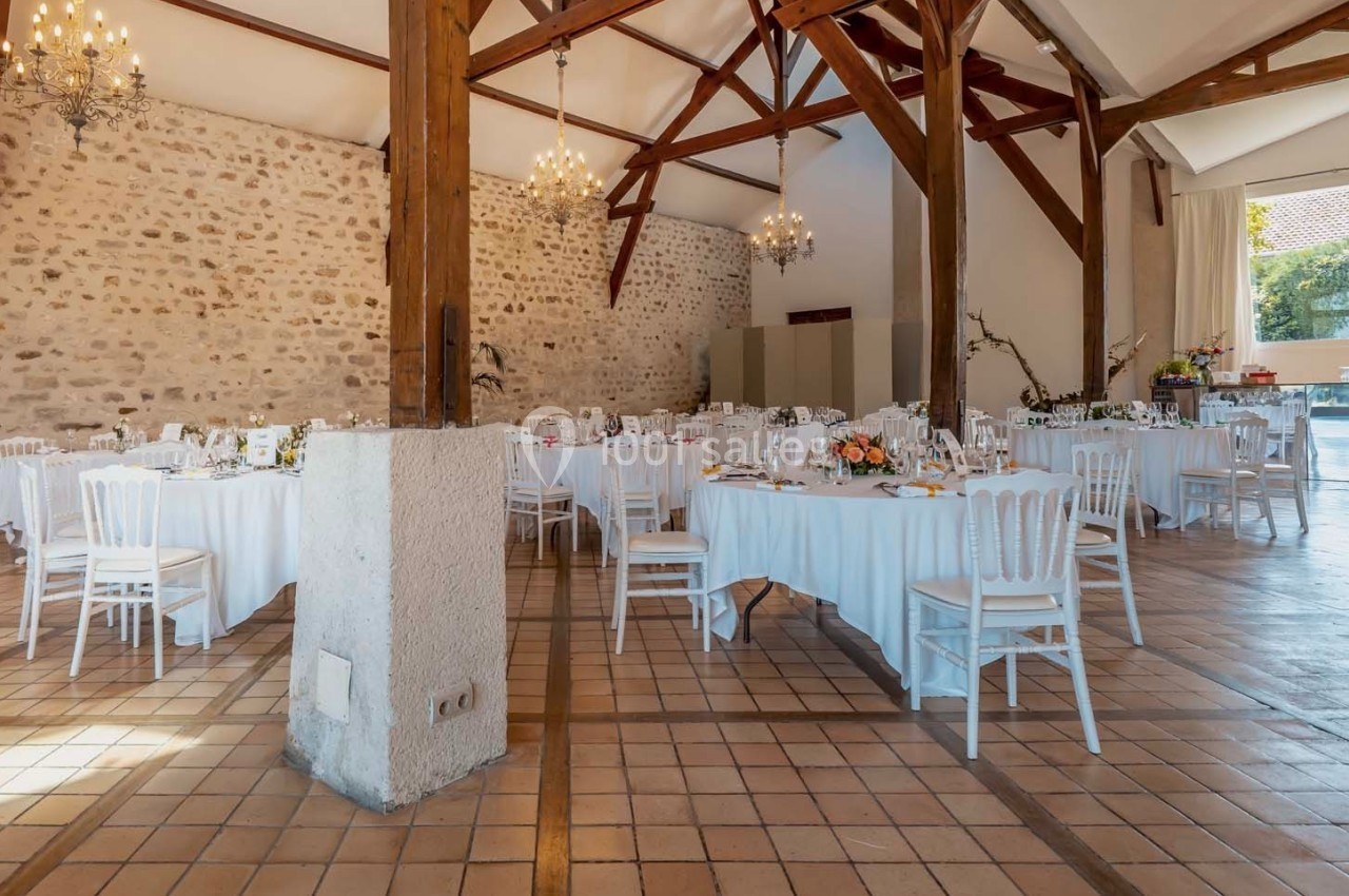 Salle de réception lumineuse avec murs en pierre, poutres apparentes, tables rondes dressées et chaises blanches.