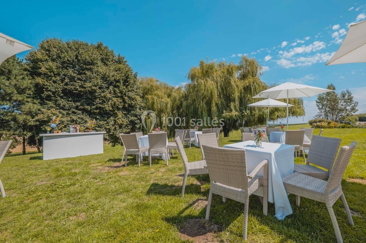 Tables et chaises en rotin disposées sur une pelouse, avec parasols blancs et arbres en arrière-plan par temps ensoleillé.