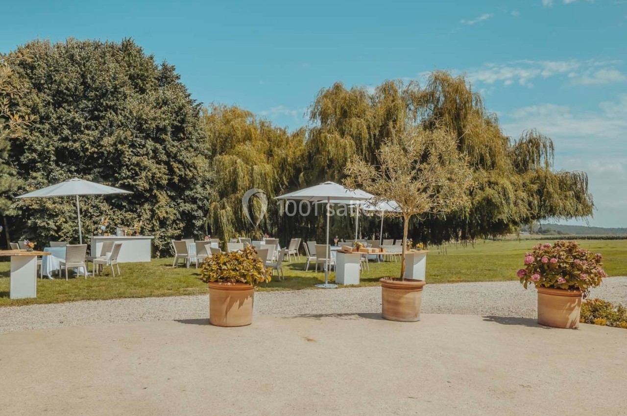 Terrasse extérieure avec tables et chaises blanches sous des parasols, entourée d'arbres et de plantes en pot.