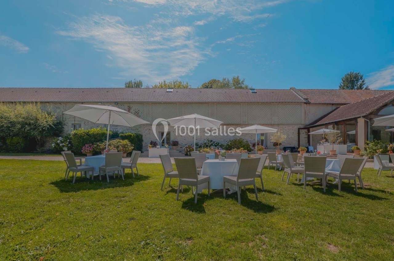 Tables et chaises disposées sur une pelouse avec parasols, devant un bâtiment en pierre sous un ciel dégagé.