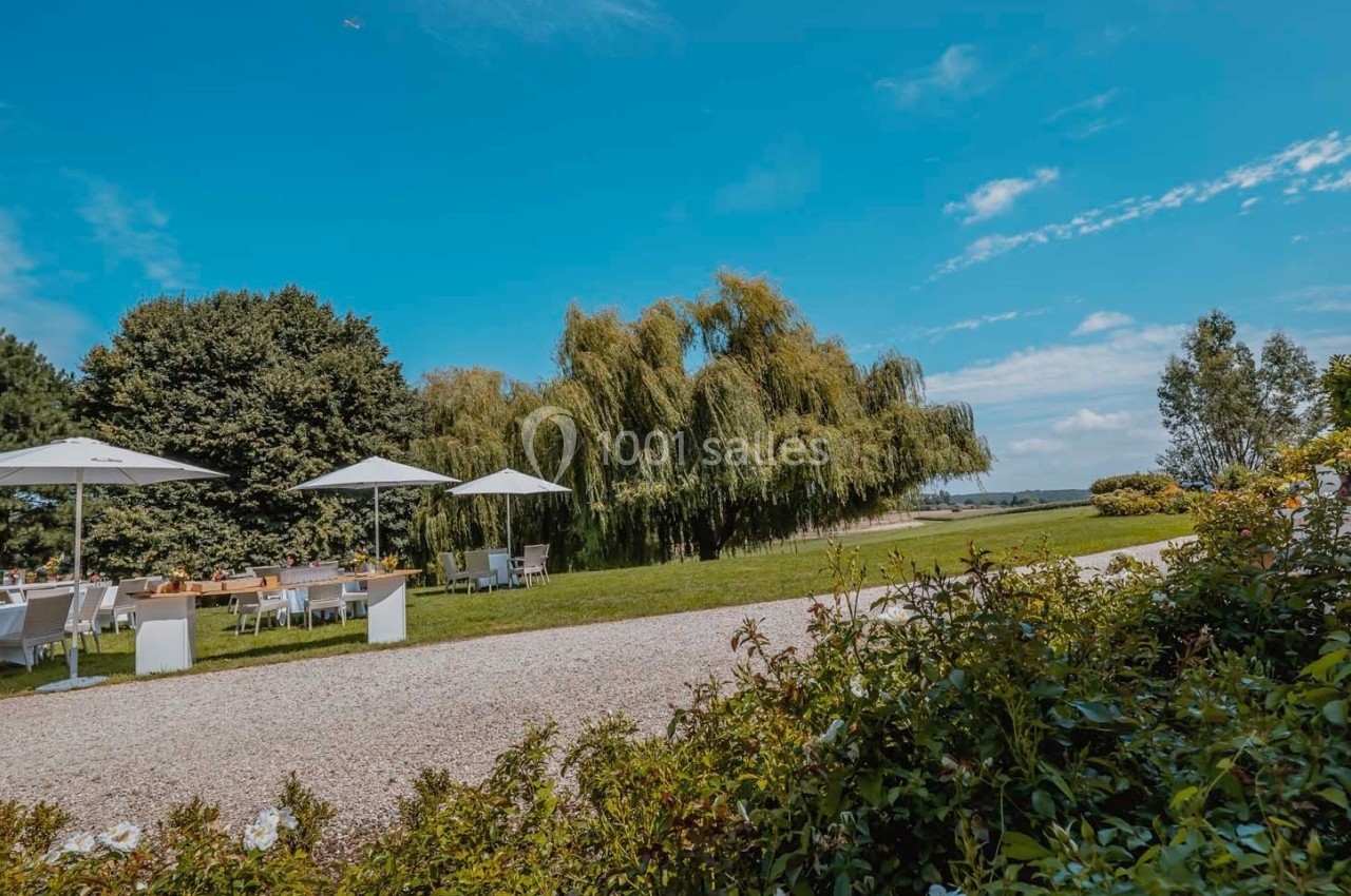 Espace extérieur avec tables et parasols près d'arbres et d'un chemin, sous un ciel bleu dégagé.