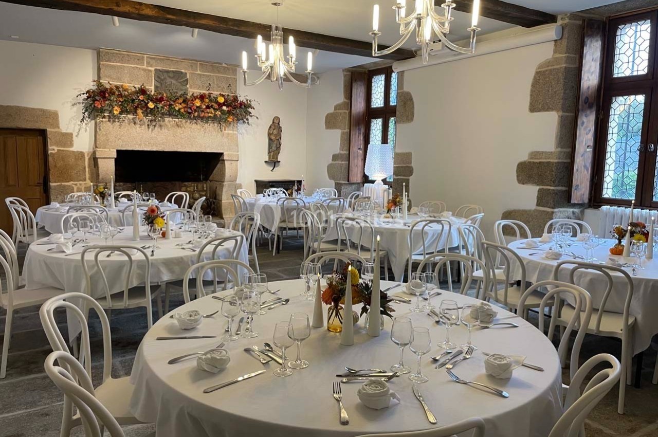 Salle de réception avec tables rondes dressées, décor automnal et cheminée en pierre ornée de fleurs.