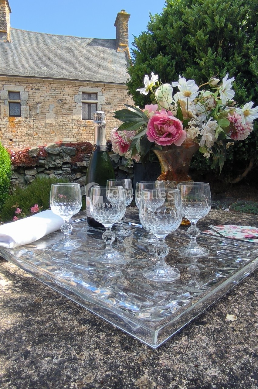 Plateau en verre avec flûtes à champagne, bouteille, fleurs et serviette, posé sur une table en extérieur près d'une maison…