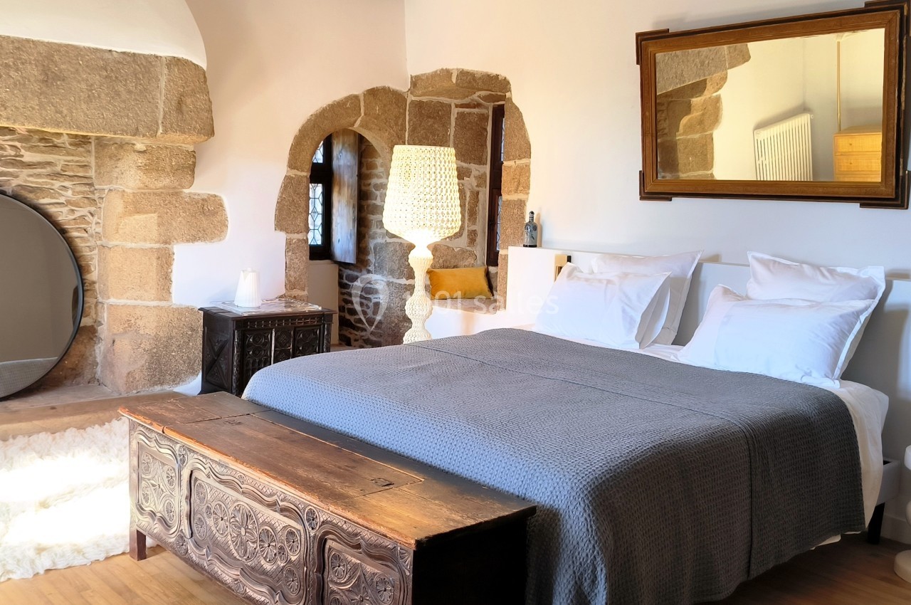 Chambre lumineuse avec lit double, murs en pierre, miroir rond, coffre en bois et décoration épurée.