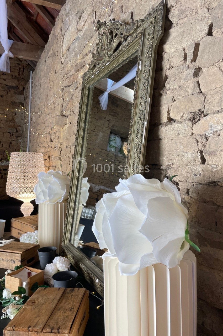 Grand miroir doré encadré, posé contre un mur en pierre, entouré de décorations et fleurs blanches.