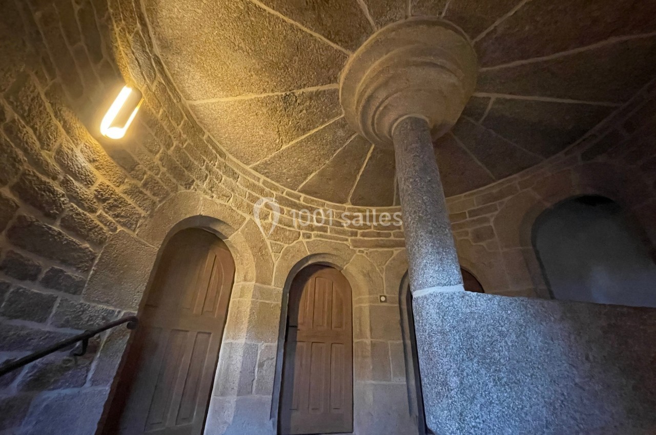 Escalier en pierre avec une colonne centrale, entouré de portes en bois et éclairé par une lampe murale.
