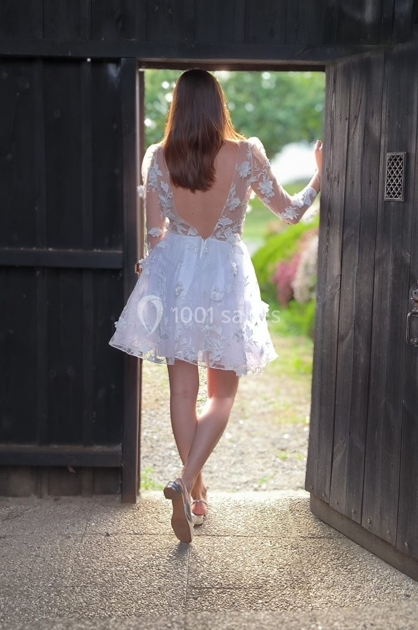 Femme en robe blanche à dos nu, sortant par une porte en bois ouverte vers un jardin lumineux.