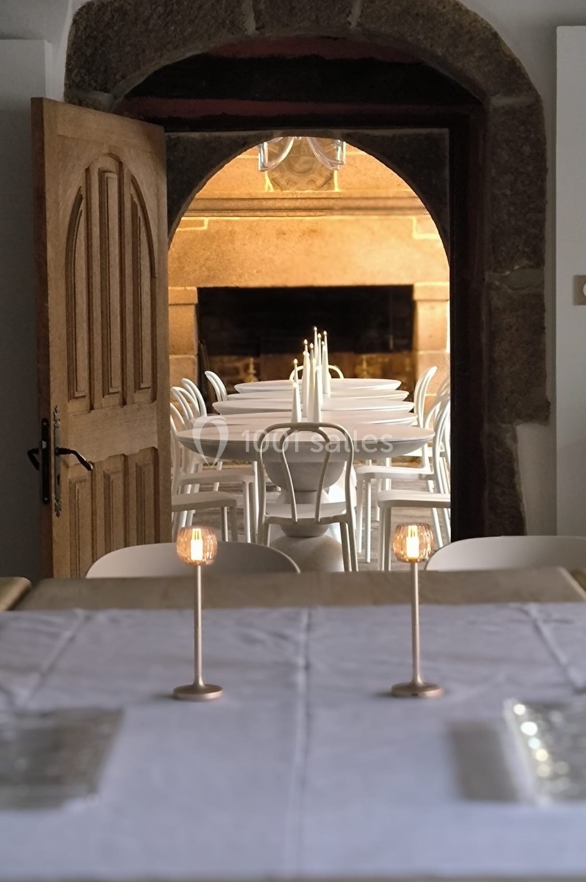 Salle à manger avec une grande table dressée, chandeliers et chaises blanches, vue sur une pièce voisine en pierre.