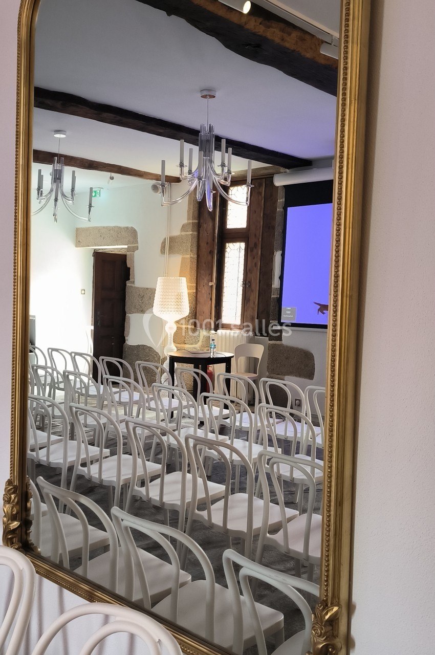 Miroir doré reflétant une salle de conférence avec des chaises blanches alignées et un écran de projection allumé.
