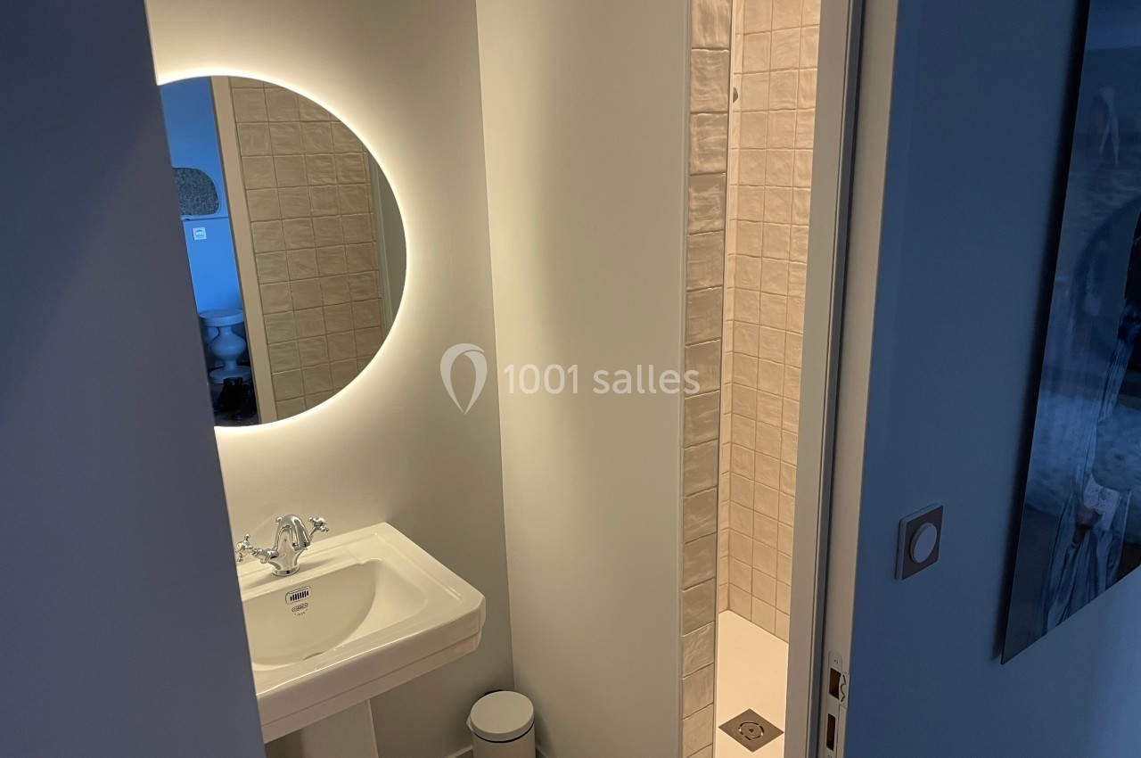 Lavabo blanc avec miroir rond éclairé, situé près d'une douche à carreaux clairs dans une petite salle de bain.