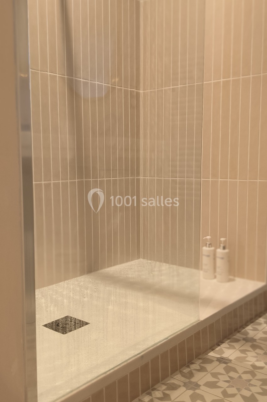Douche moderne avec parois en verre, carrelage beige vertical et sol à motifs géométriques, deux flacons posés au fond.