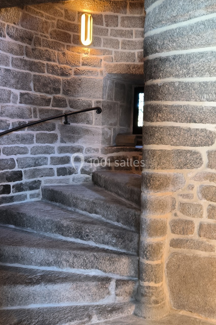 Escalier en pierre courbé avec une rampe en métal, éclairé par une applique murale et une petite fenêtre.