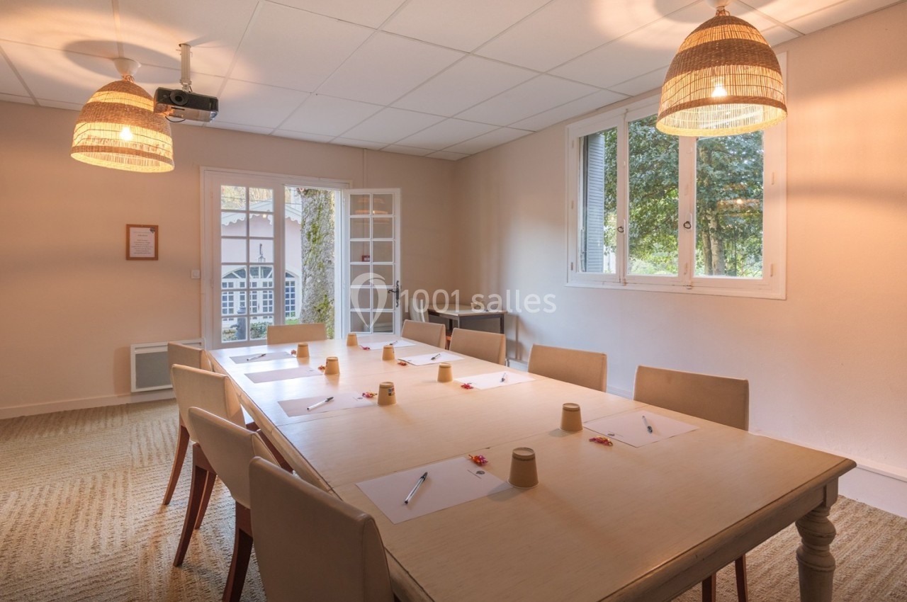 Salle de réunion lumineuse avec une grande table rectangulaire, des chaises, des documents et des stylos disposés.