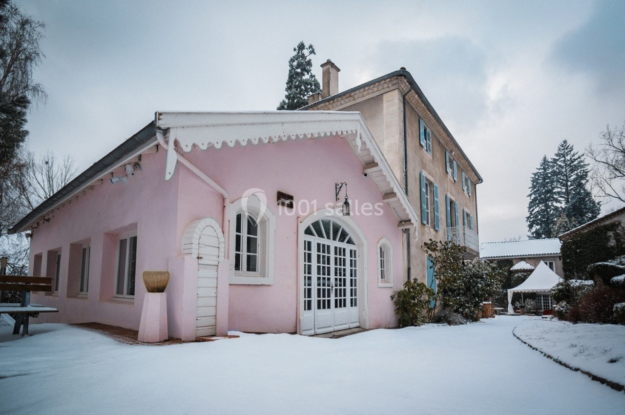 Bâtiment rose et maison beige adjacente entourés de neige, avec arbres en arrière-plan sous un ciel nuageux.