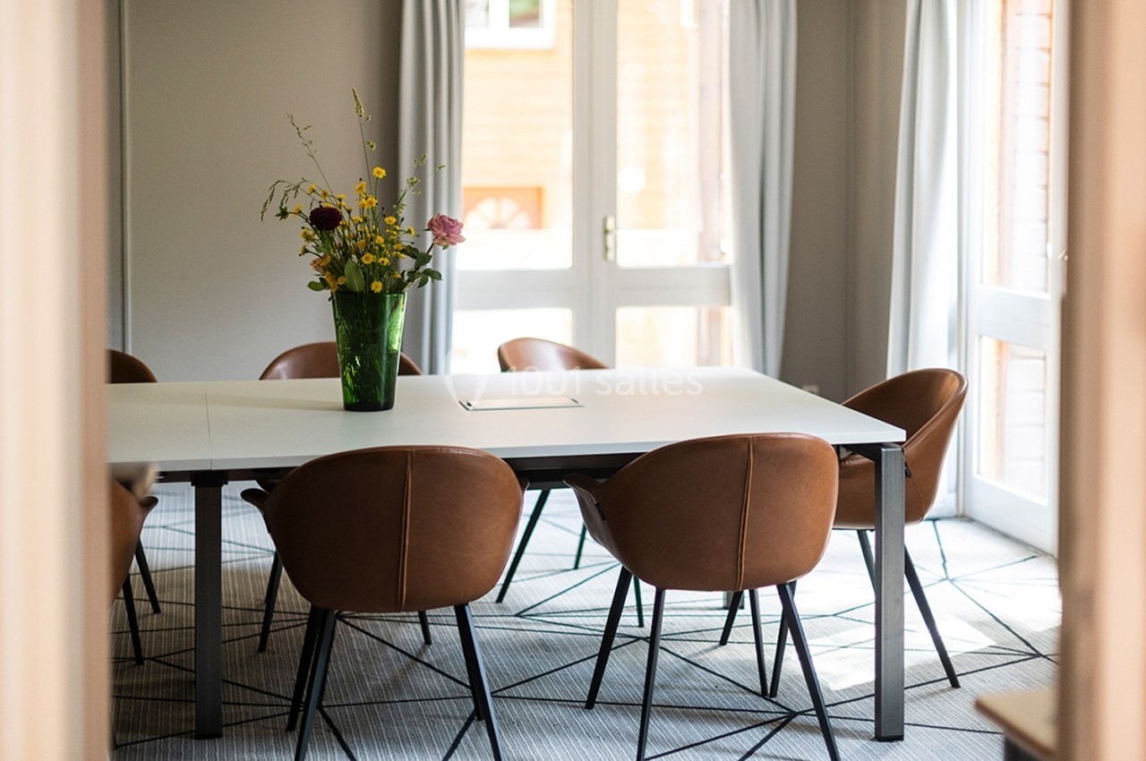 Salle lumineuse avec table blanche, chaises en cuir marron et vase de fleurs colorées au centre.