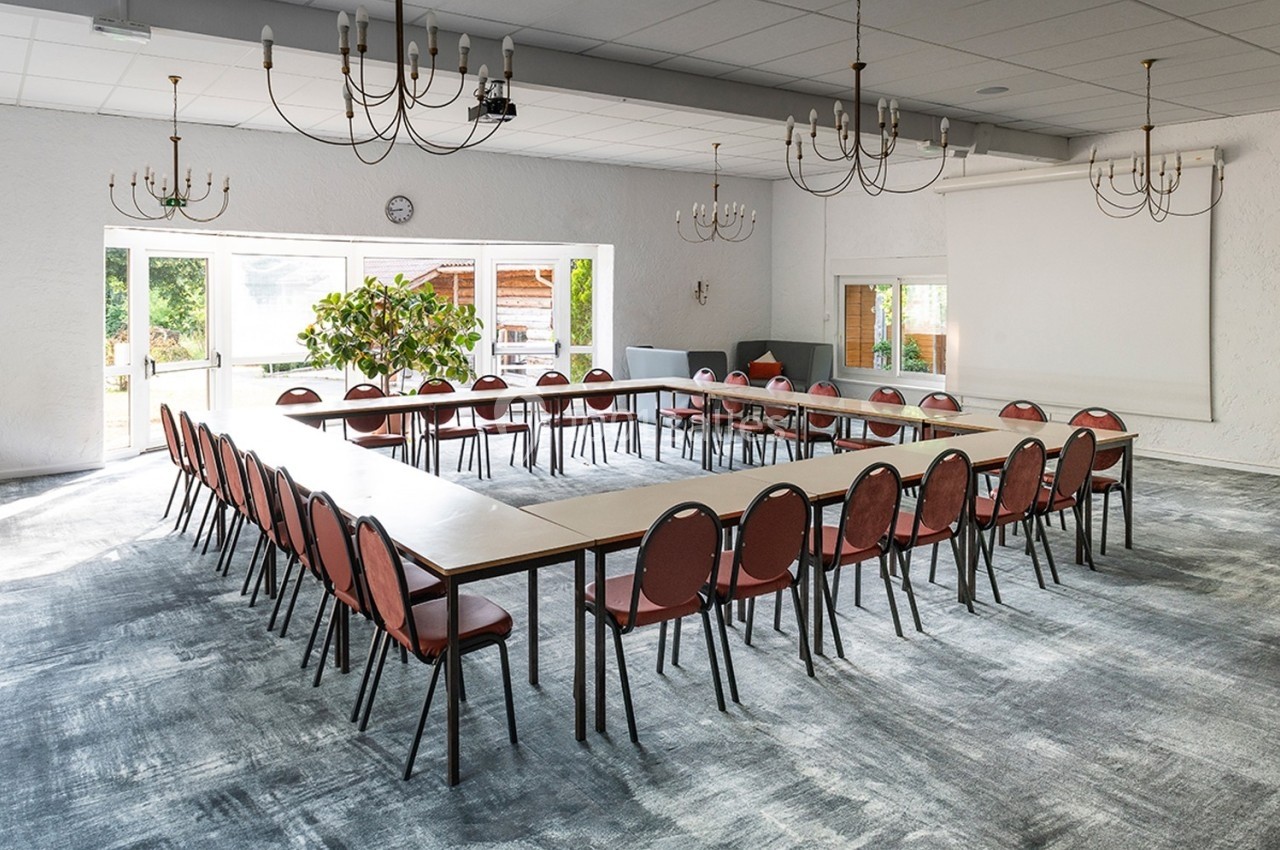 Salle de réunion lumineuse avec des tables disposées en U, entourées de chaises rouges et éclairée par des fenêtres.