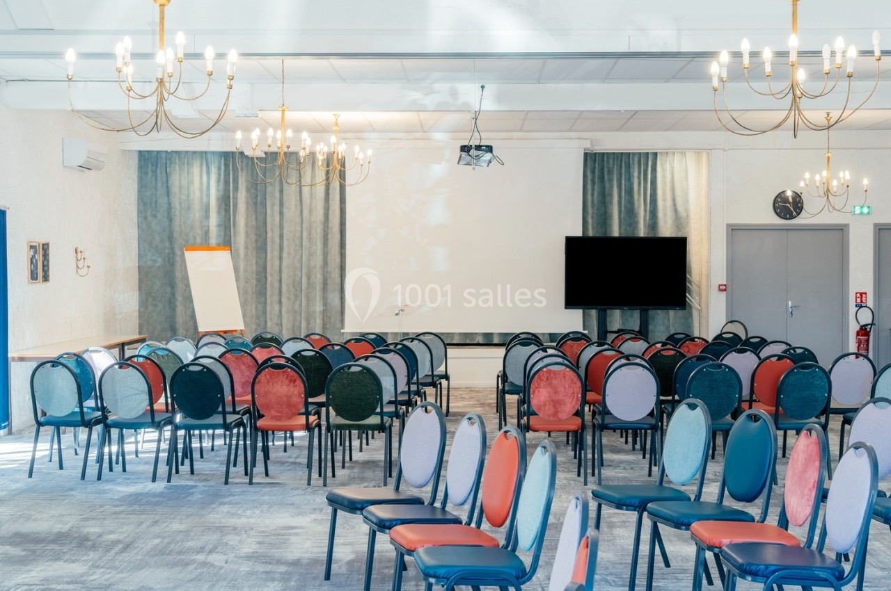 Salle de conférence lumineuse avec des rangées de chaises colorées, un écran de projection et un tableau blanc.