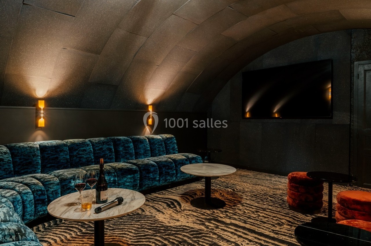 Salon cosy avec canapé en velours bleu, tables basses, éclairage tamisé et grand écran mural.