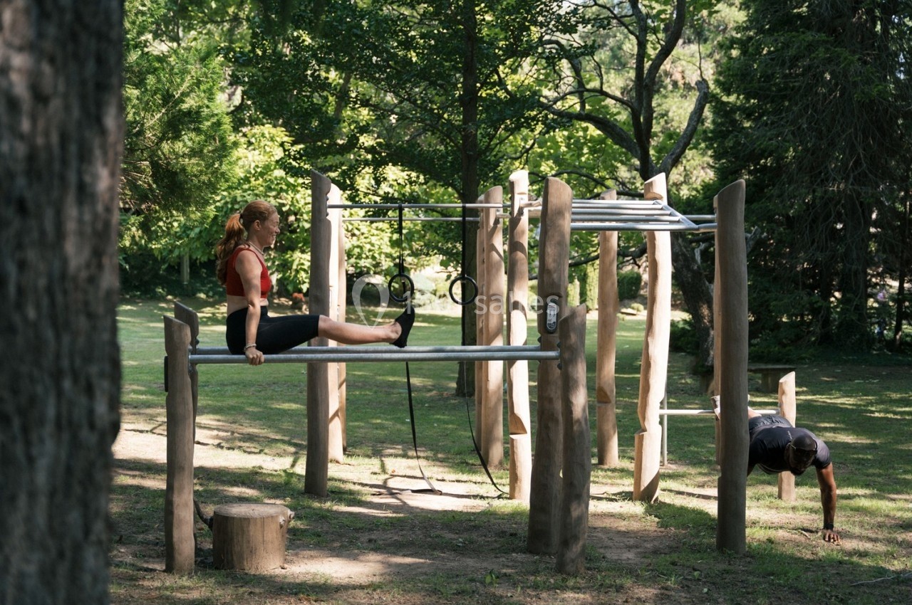 Une femme fait des exercices sur des barres parallèles dans un parc, entourée d'arbres et d'équipements sportifs.