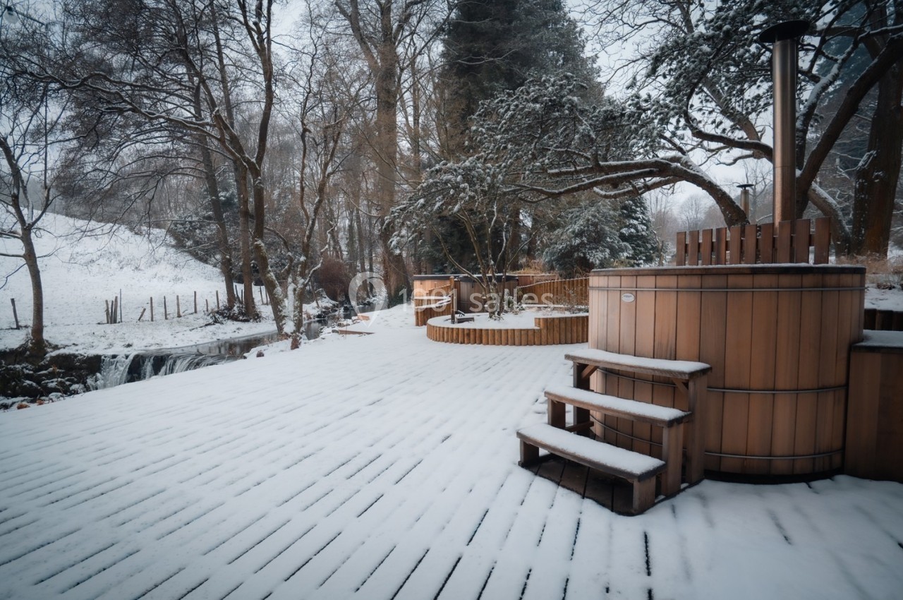 Terrasse en bois enneigée avec bains nordiques entourés d'arbres et d'un paysage hivernal.