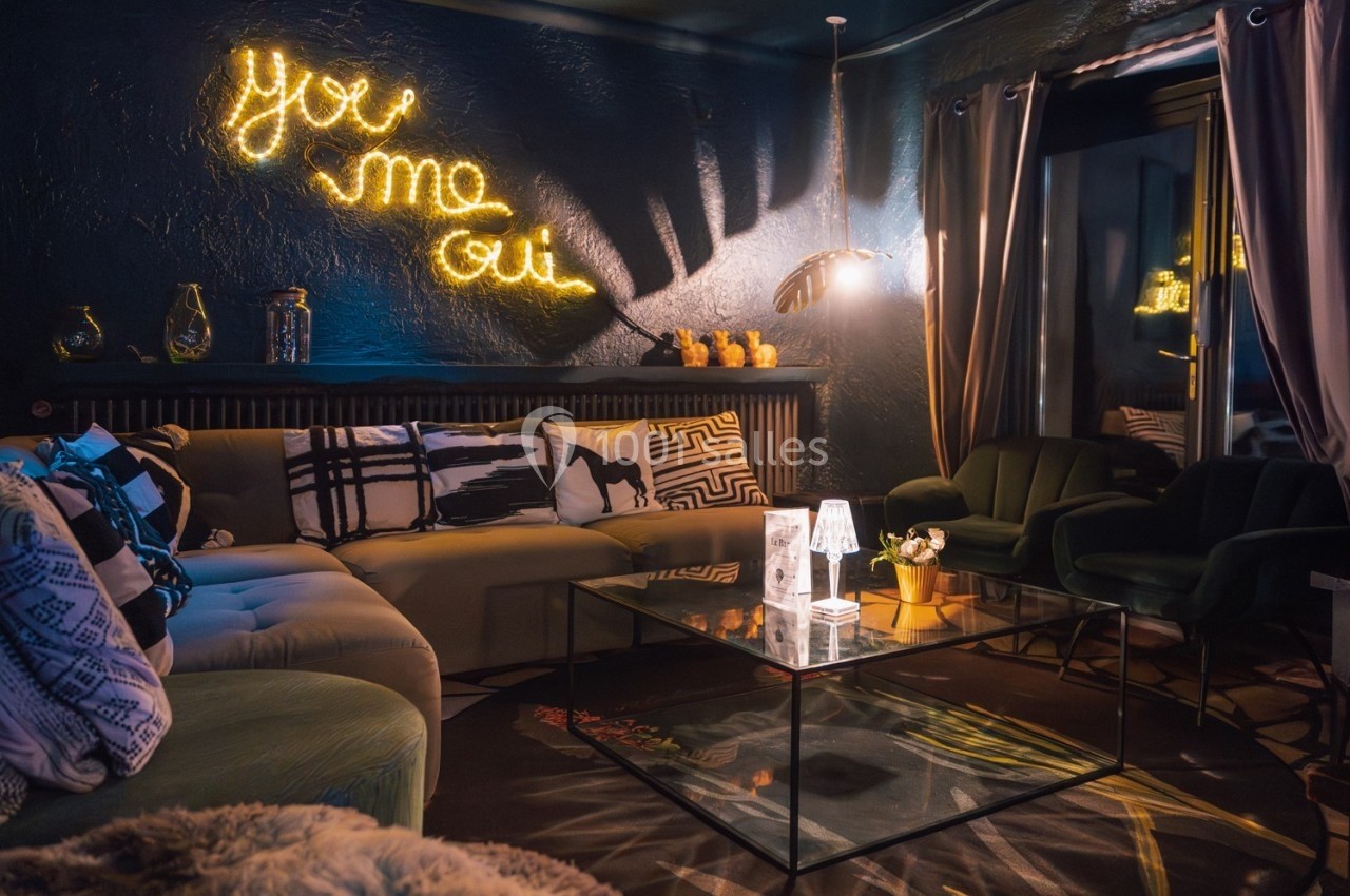 Salon cosy avec canapé, fauteuils, table basse en verre, éclairage tamisé et néon mural ’You me oui’.