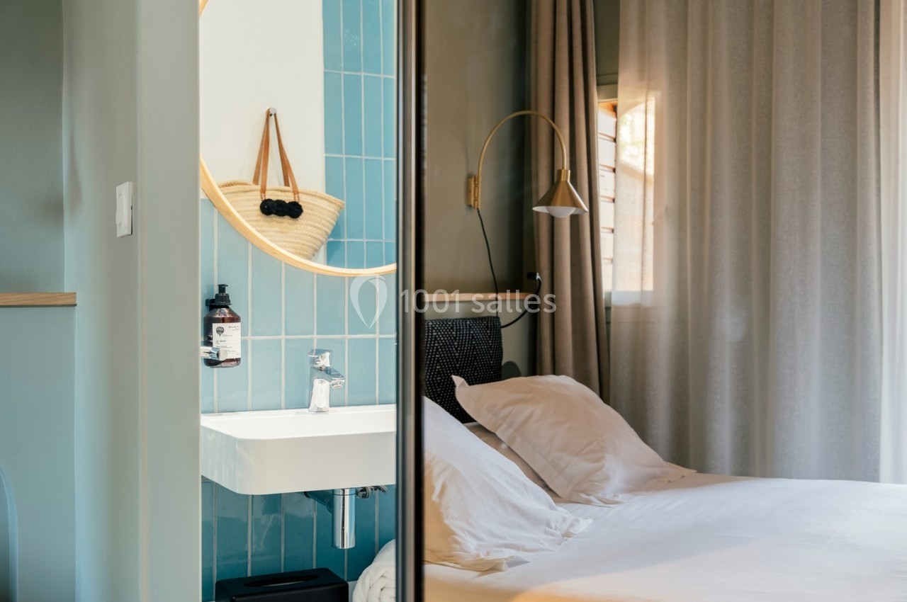 Vue d'une chambre lumineuse avec un lit blanc, un miroir rond au-dessus d'un lavabo et des carreaux bleus.