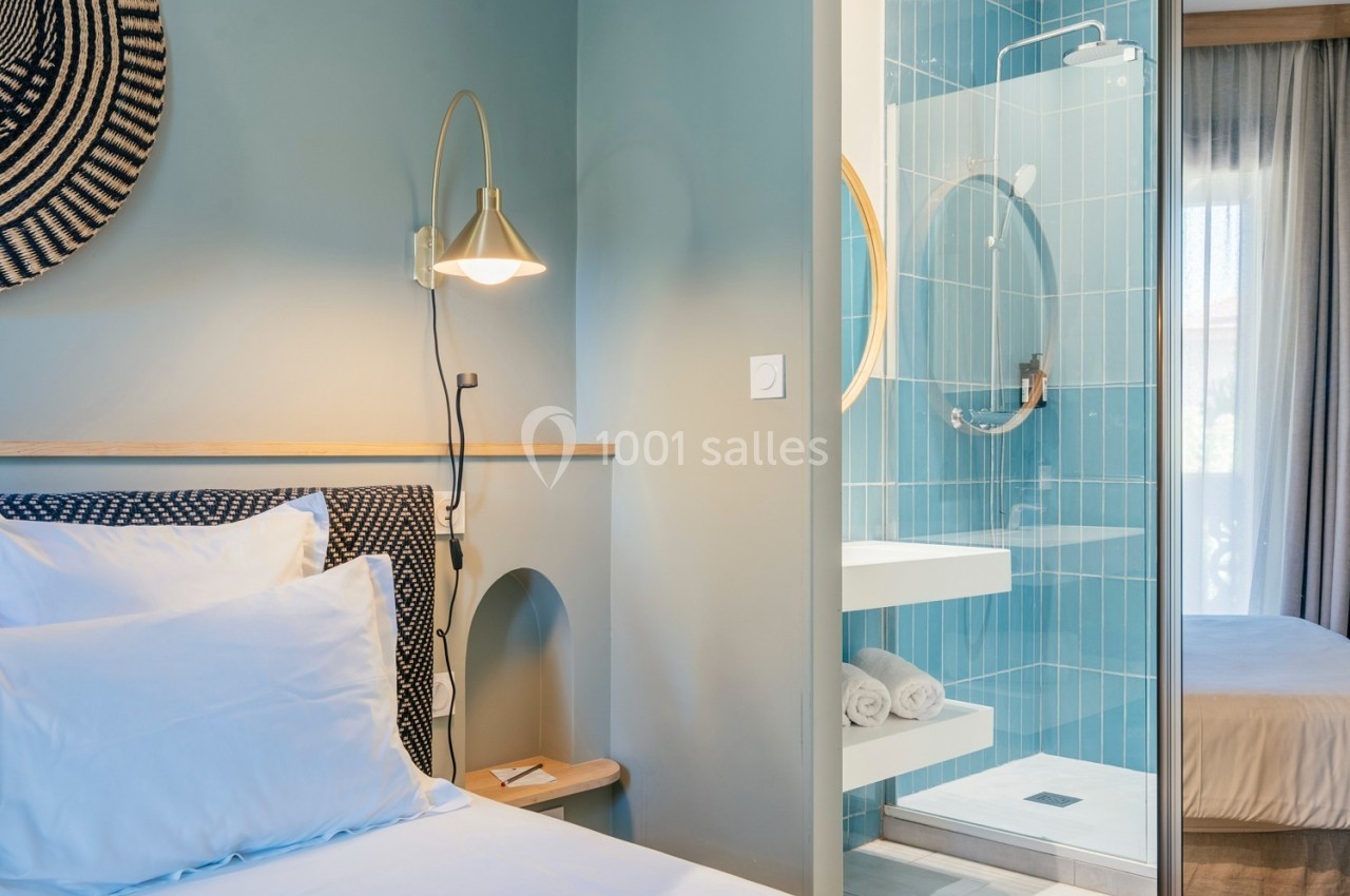 Chambre lumineuse avec lit double, mur bleu, lampe murale et salle de bain attenante aux carreaux bleus.