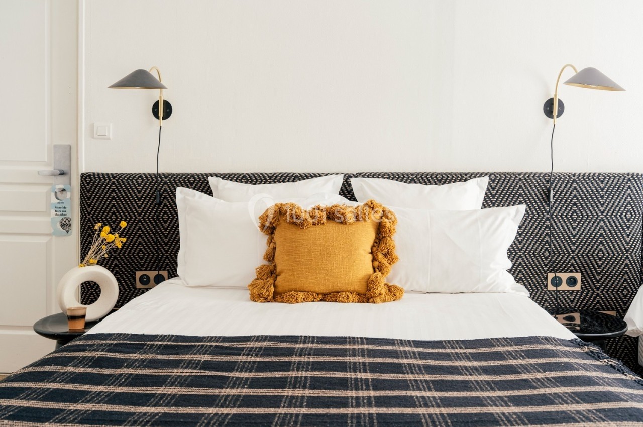 Lit double avec coussins blancs et un coussin décoratif jaune, encadré par deux appliques murales noires.