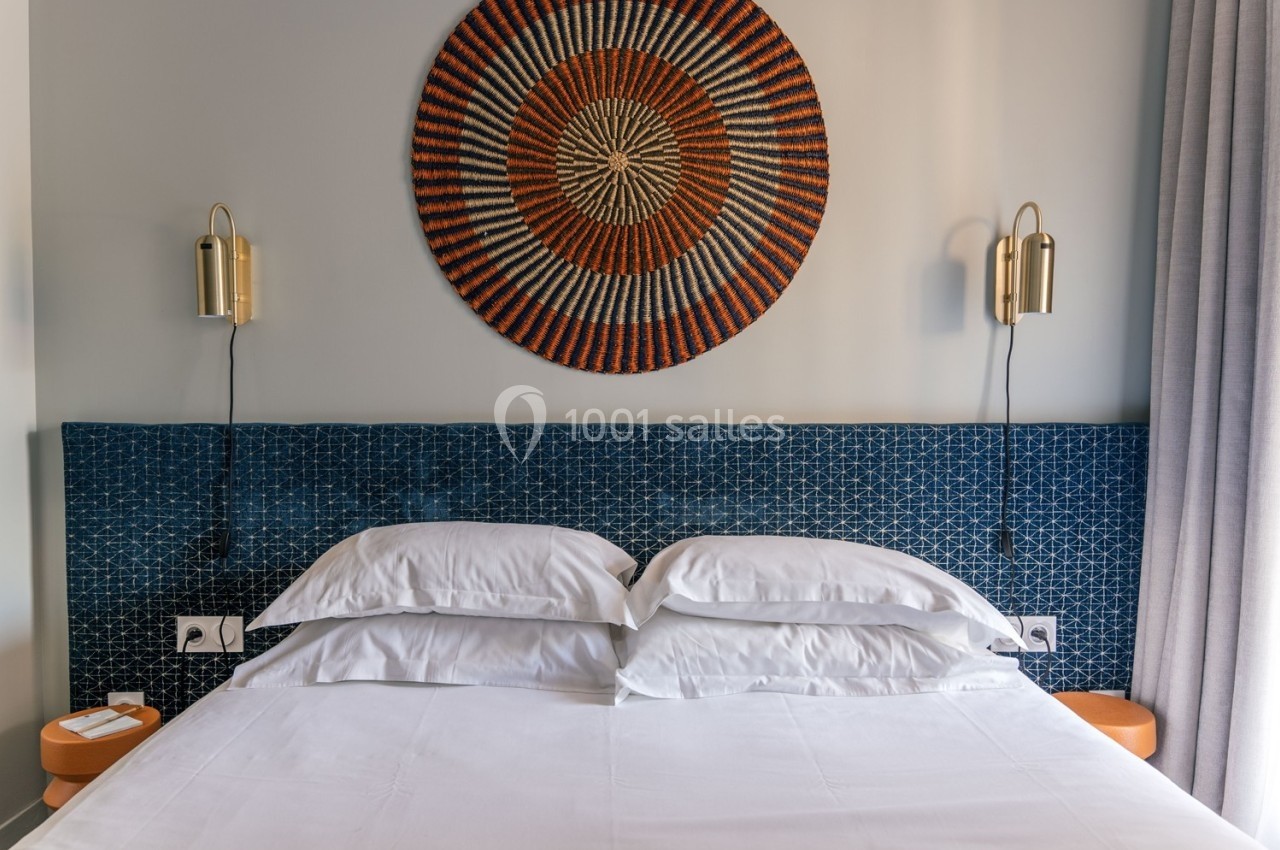 Lit double avec tête de lit bleue texturée, décoré d'un grand panier mural coloré et éclairé par deux appliques dorées.