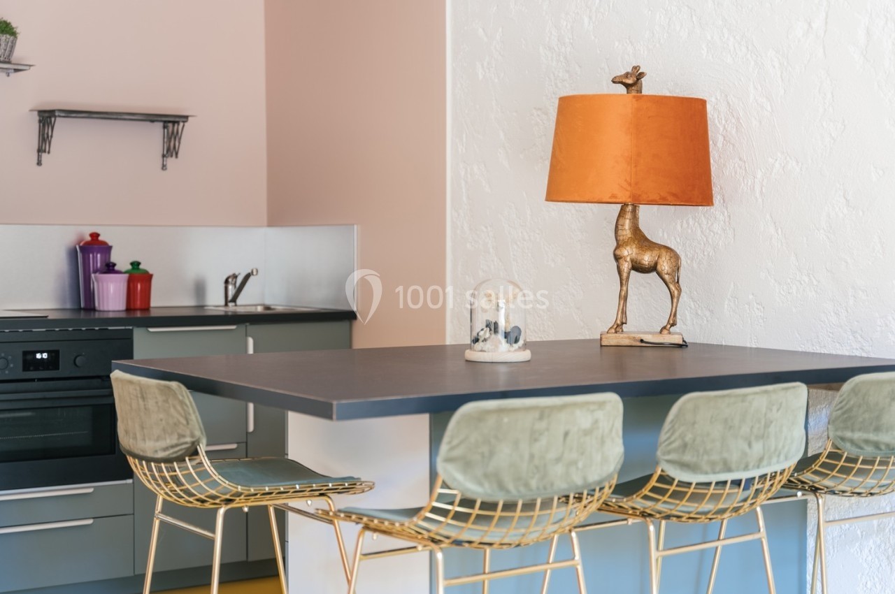 Cuisine moderne avec un plan de travail, des tabourets dorés, une lampe décorative en forme de girafe et des étagères…