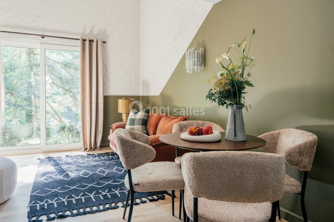 Salle de séjour lumineuse avec table ronde, chaises en tissu, canapé orange, tapis bleu et grande baie vitrée.