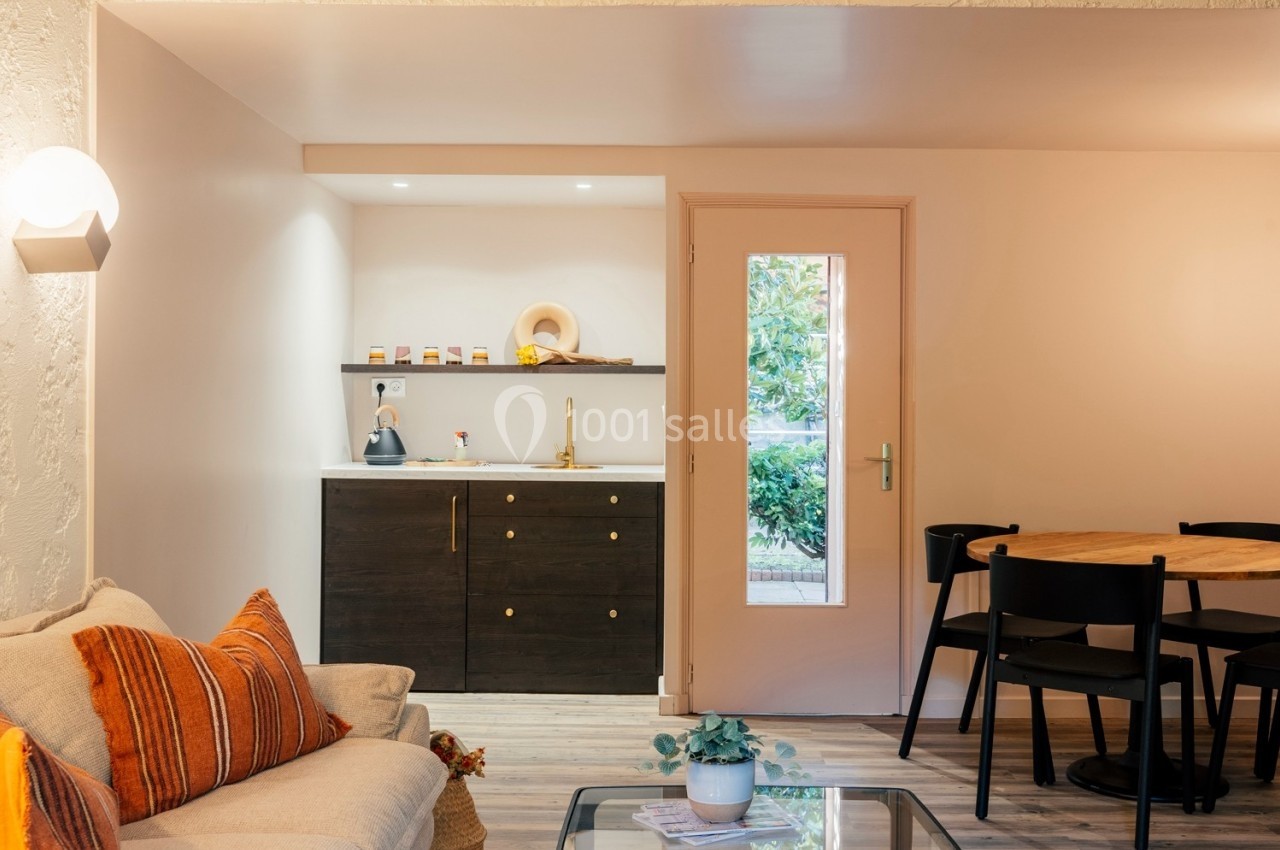 Coin salon avec canapé, table basse et cuisine ouverte équipée, éclairé par une porte vitrée donnant sur l'extérieur.