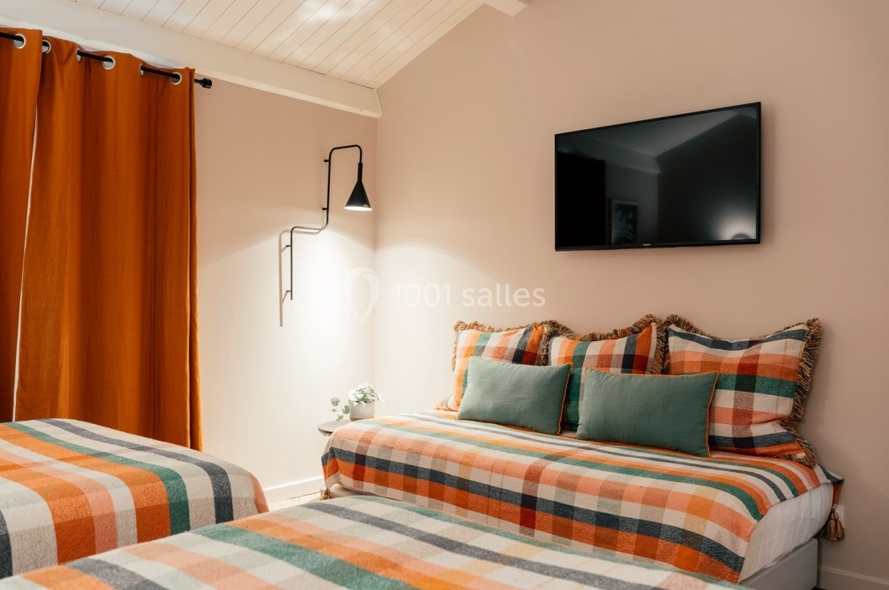 Chambre avec lits et coussins à motifs colorés, rideaux orange, mur beige, téléviseur mural et éclairage moderne.