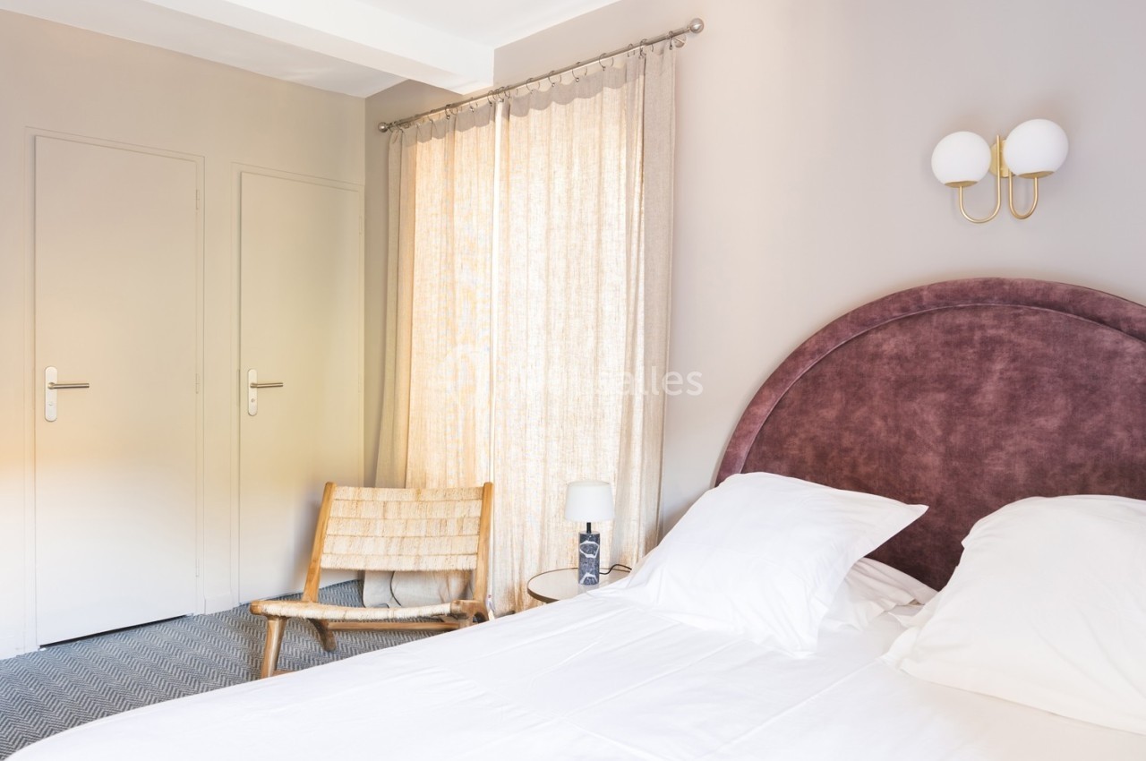 Chambre lumineuse avec lit à tête de lit violette, chaise en bois, rideaux beiges et applique murale blanche.