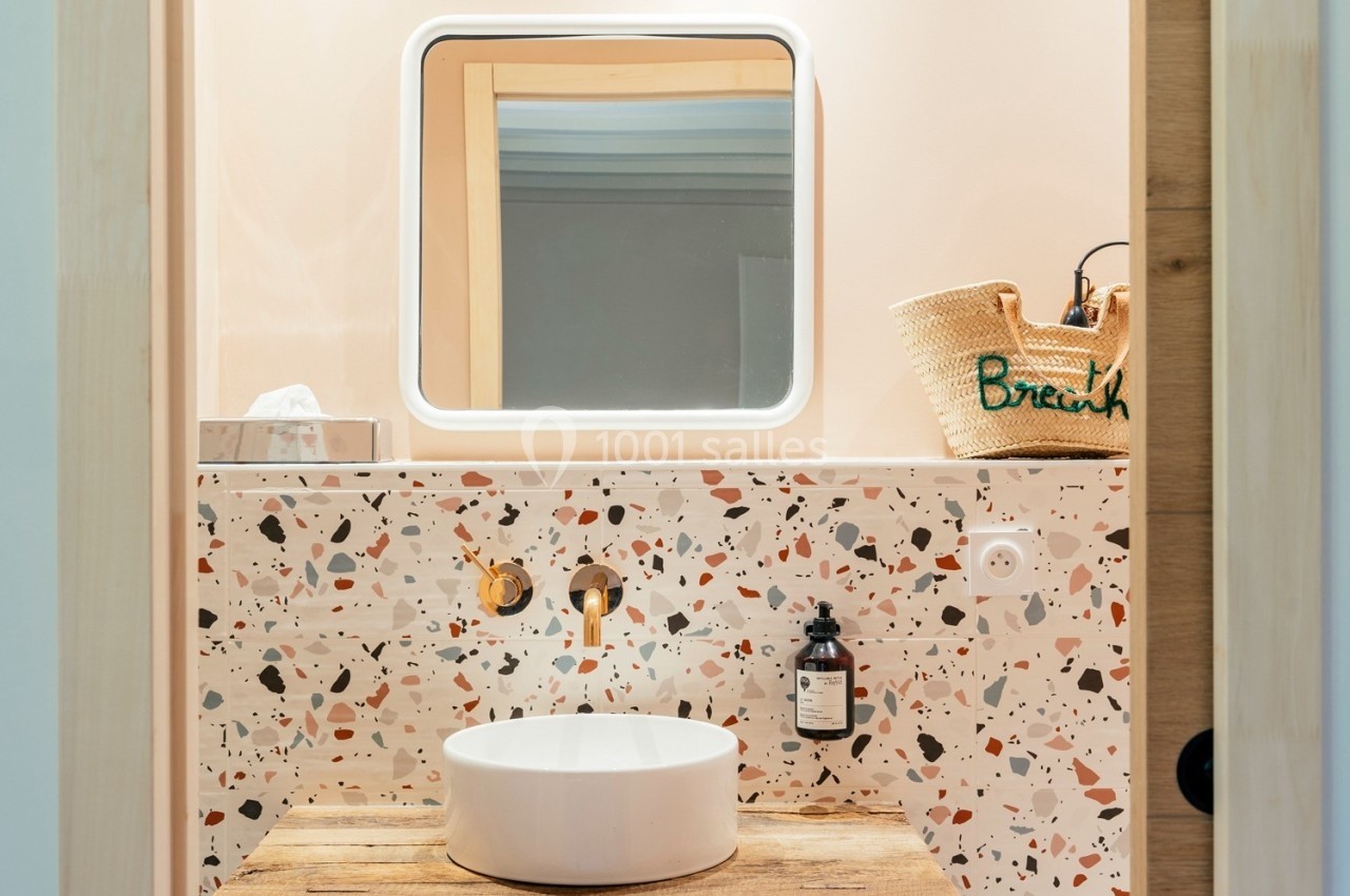 Lavabo rond sur un plan en terrazzo coloré, avec miroir rétroéclairé et panier en osier posé sur le côté.