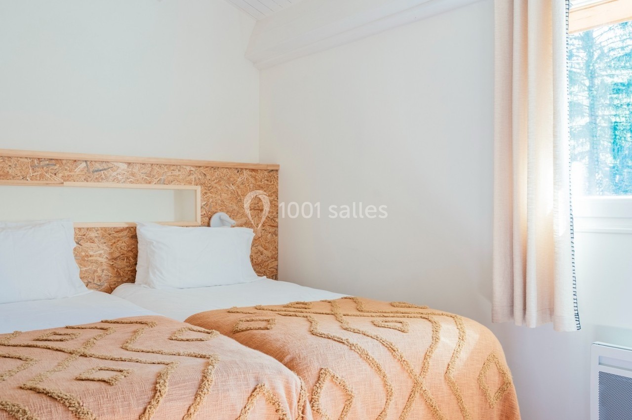Deux lits simples avec couvertures orange et coussins blancs dans une chambre lumineuse avec fenêtre et rideaux beiges.