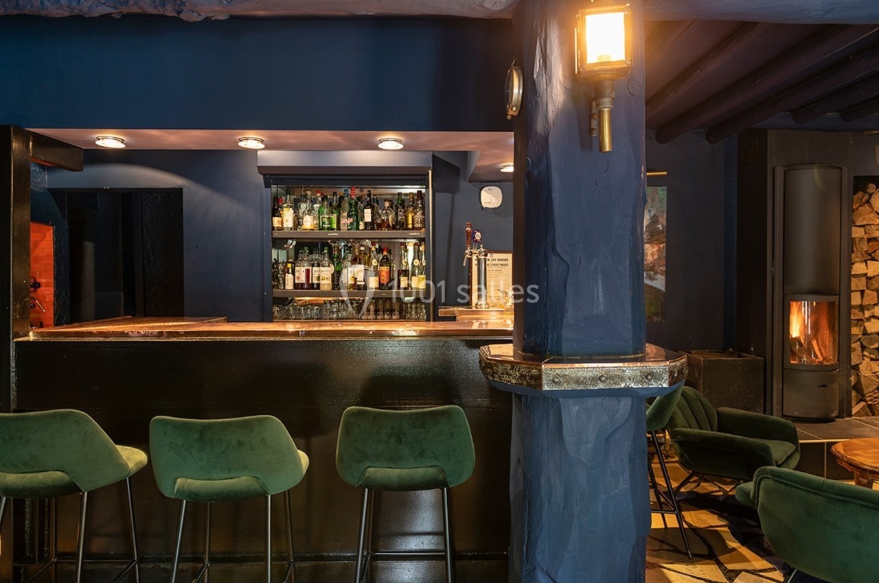 Bar avec comptoir en bois sombre, tabourets verts, étagères de bouteilles et éclairage chaleureux dans une ambiance cosy.