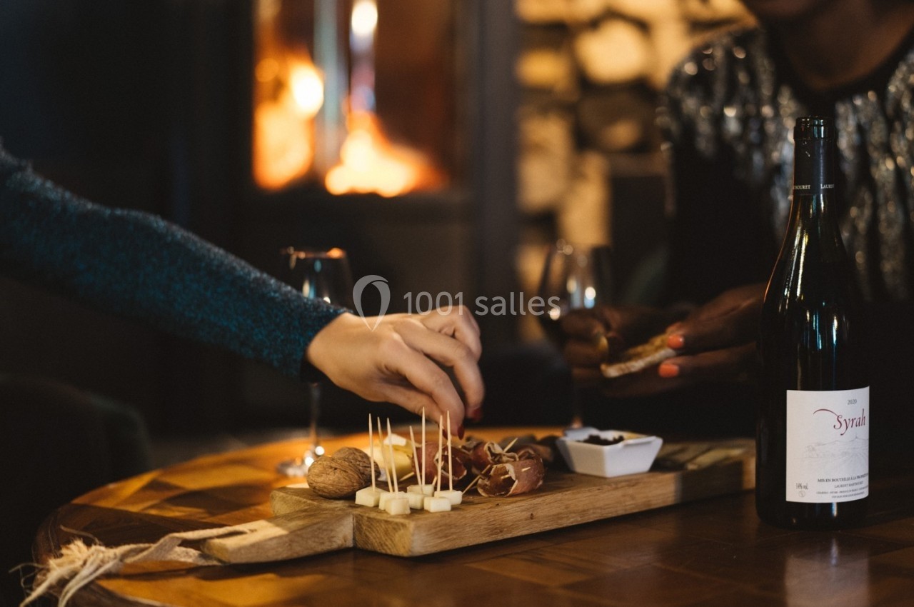 Plateau de fromages et noix sur une table en bois, avec des mains servant et un feu de cheminée en arrière-plan.