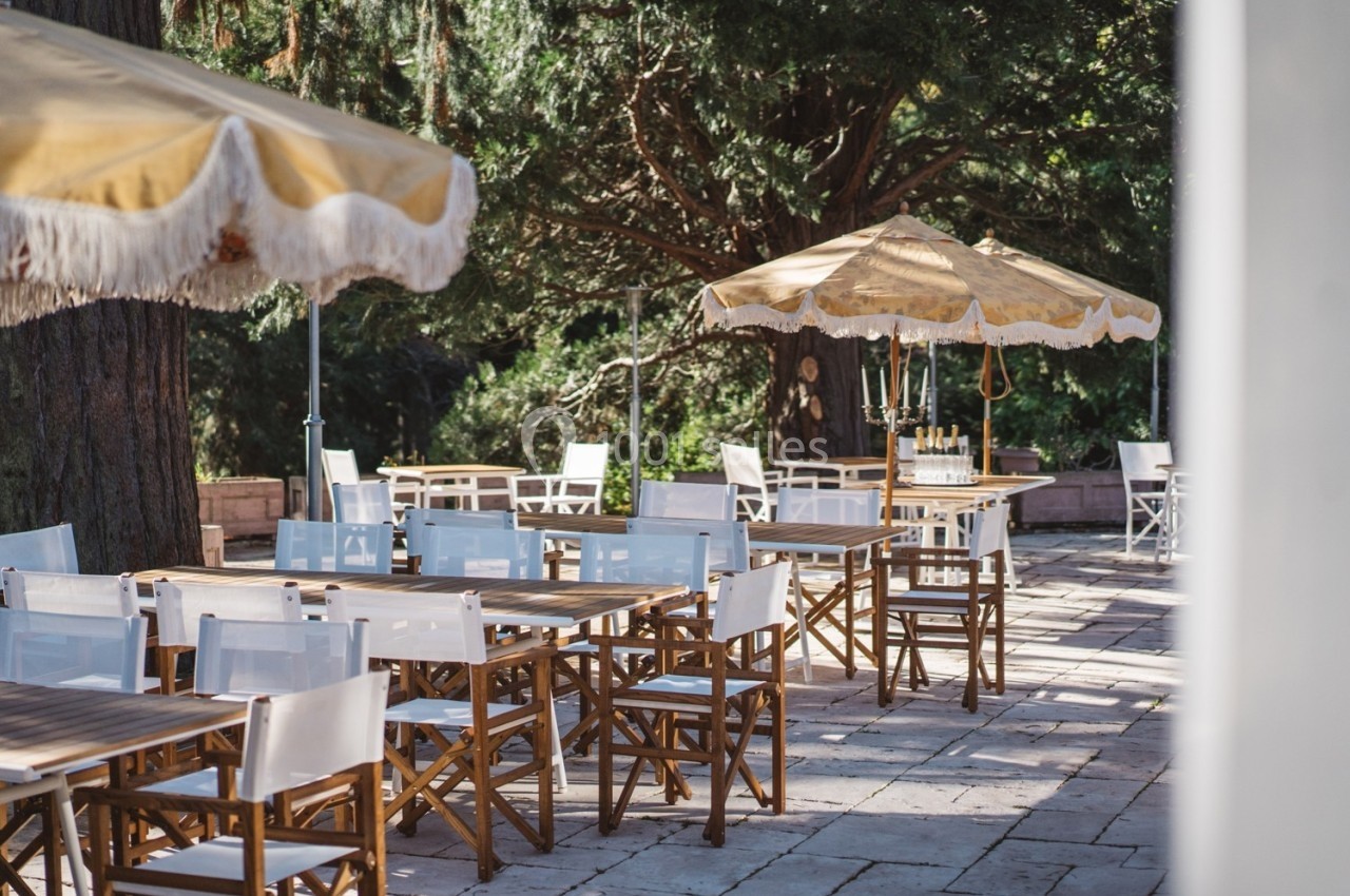 Terrasse extérieure avec tables et chaises en bois, parasols beige et végétation en arrière-plan.