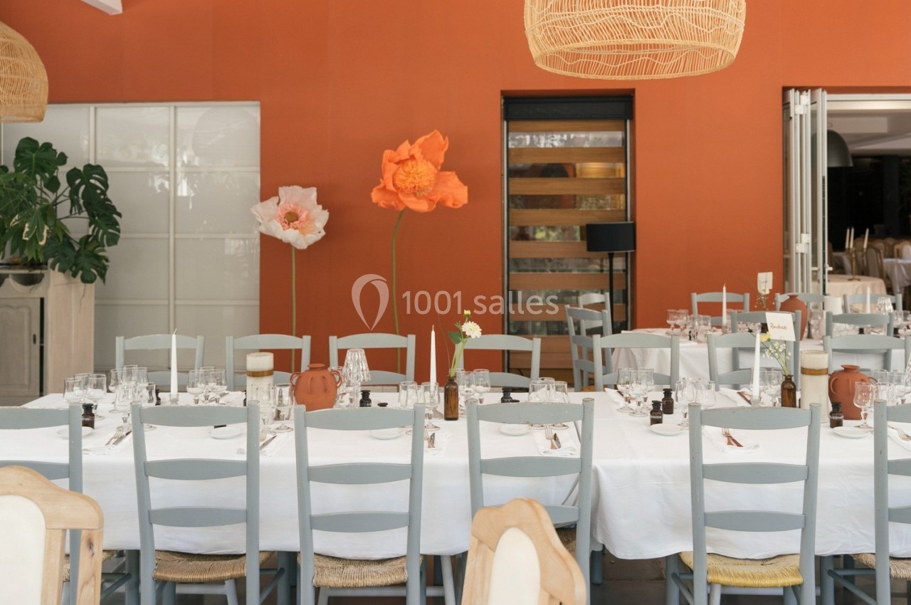 Salle de réception avec tables dressées, nappes blanches, chaises grises et décorations florales devant un mur orange.