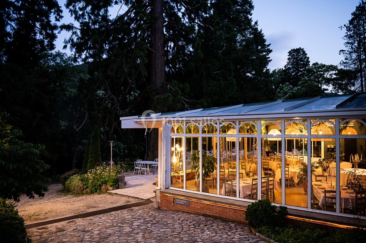 Salle de réception vitrée éclairée en soirée, entourée de végétation et d'une allée pavée.
